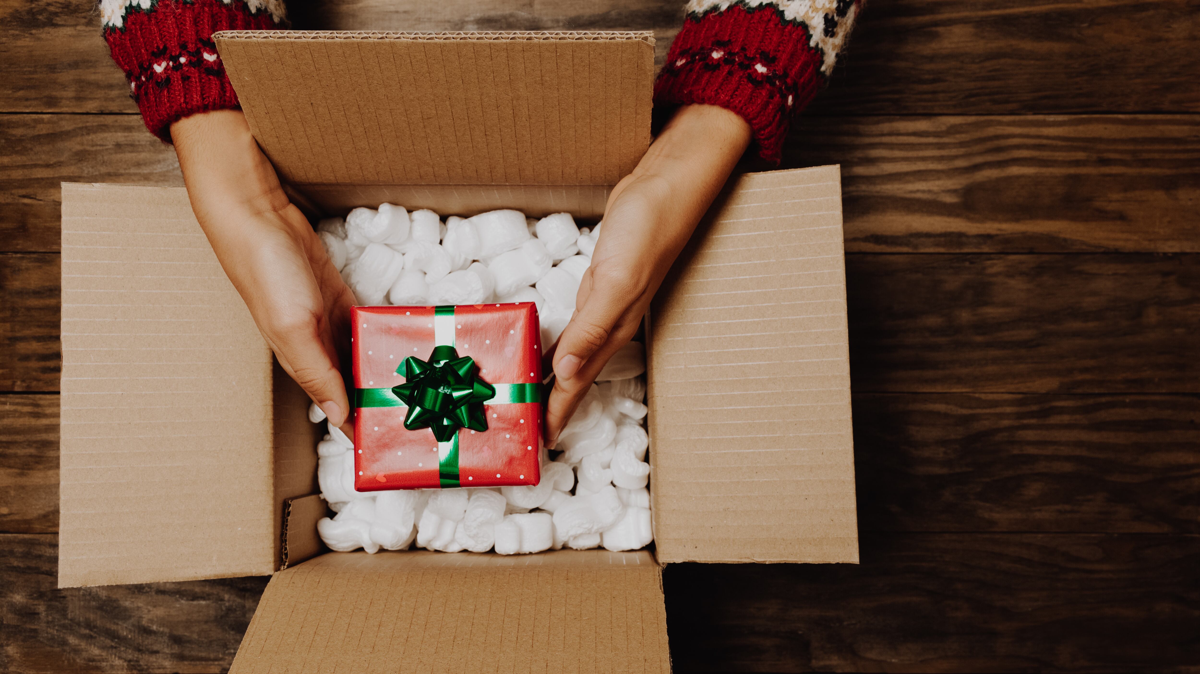 Female hands with a winter sweater packing or unpacking a Christmas gift from a cardboard box on a rustic wooden table.